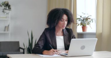 Evden online danışmanlık yapan odaklanmış genç bayan avukat, müşterilerle iletişim kurmak için modern bilgisayarın webcam 'ini kullanıyor, video görüşmeleri ile müşteriyle konuşuyor, kağıda not alıyor, notlar alıyor.