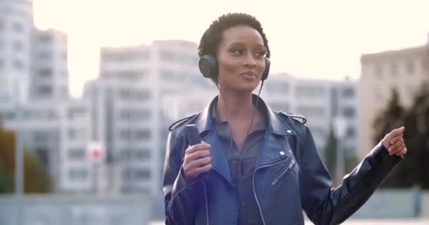 Happy African American nastolatka dziewczyna nosi skórzaną kurtkę i słuchawki, trzyma smartfona w rękach, tańczy do muzyki na ulicy. Stylowe mieszane rasy kobieta przenosi się na zewnątrz do dźwięków słuchawek telefonu — Wideo stockowe