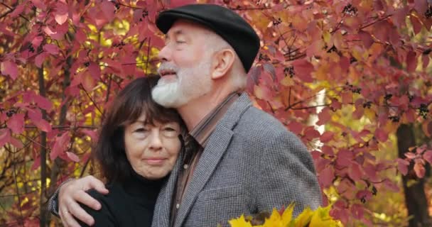 Primer plano pareja de ancianos, marido barbudo con el pelo gris abrazar a la triste esposa con ramo de hojas de otoño, consolador regocijo bromas sonriendo y riendo. Relaciones románticas caminando ciudadanos mayores. — Vídeos de Stock