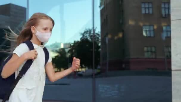 Schoolgirl in dress with backpack wears medical mask, pandemic time, protection against corona virus, prevention infection. Little girl running from school class meeting Mom, parent hugging daughter — Stock Video