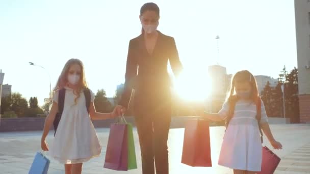 La familia joven camina calle abajo, usando máscaras médicas protectoras, llevando bolsas de regalos. Mamá de negocios recoge a dos hijas después de la escuela durante la epidemia de virus pandémico, ir con los niños tomados de la mano — Vídeo de stock