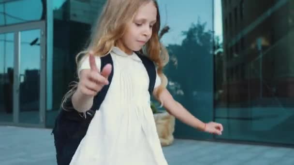 Lonely active little girl with golden hair wears dress and backpack, walks along city street, runs jumps joyfully against background of glass building, rushes elementary school for first grade lesson — Stock Video