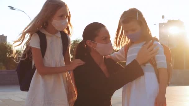 Jeune mère brune accompagne deux filles à l'école pour la leçon, les gens s'assoient dehors sous des masques médicaux protecteurs, premier jour de septembre, étude pendant la pandémie de virus, risque d'infection proche famille — Video