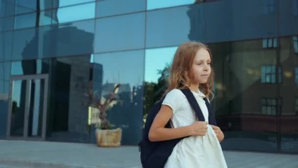 Estudante bonito com cabelo dourado carrega mochila grande, caminha para casa da escola através da cidade, salta feliz, corre ao longo da estrada. As notas elementares dos alunos vão para a aula para colegas de classe, sorrindo — Vídeo de Stock