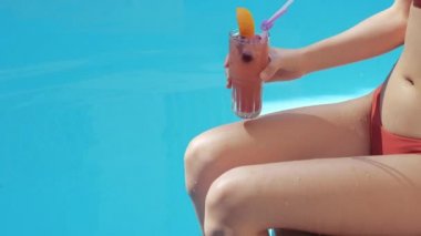Asyalı bir esmer kadının yakın plan fotoğrafı kırmızı şık mayolu, havuz kenarında oturuyor, ayaklarını serin suya daldırıyor, hayal kuruyor, barın yakınında sağlıklı bir kokteyl içiyor, atış talimi yapıyor.