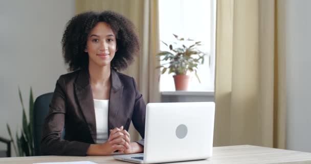 Vrouw in zakelijke stijl kledij zit aan de werktafel in kantoor kamer met laptop apparaat, kijkt vriendelijk naar de camera, glimlacht kieskeurig, schudt haar hoofd in overeenstemming, drukt steun en groeten — Stockvideo