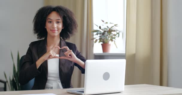 Close-up vriendelijke jonge Afro-Amerikaanse zakenmeisje zit aan bureau tafel met laptop, glimlacht tandjes, vouwt haar handen in het hart teken voor haar, concept van als, hoop, vertrouwen, vriendschap — Stockvideo