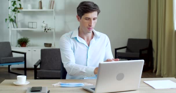 Homem engraçado senta-se na mesa, usa spray de gel desinfetante em garrafa para desinfetar as mãos lavar laptop, trata local de trabalho do vírus micróbios, coloca três máscaras médicas na cabeça dos olhos da boca, proteção corona completa — Vídeo de Stock