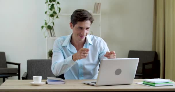 Молодой привлекательный улыбающийся мужчина сидит за столом перед ноутбуком, общается онлайн с веб-камерой на видеоконференции, флиртует дружелюбно, указывает пальцем на экран, поддерживает диалог, концепцию удаленной работы — стоковое видео
