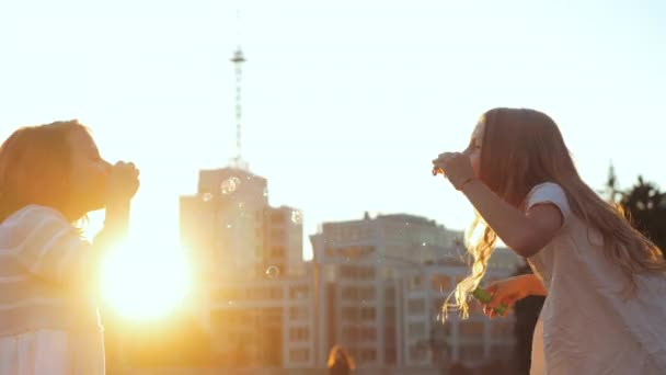 Cerca de dos niñas hermanas amigas pasar tiempo juntos al aire libre en el parque de la ciudad, soplando burbujas de jabón, globos vuelan maravillosamente en el aire al atardecer fondo de los edificios, concepto de infancia despreocupado — Vídeo de stock