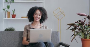 Yetişkin ırktan bir kadın iş yerindeki video konferansında dizüstü bilgisayar kullanıyor. İnternet üzerinden sorulara cevap veriyor. Afrikalı kadın vlogger el sallıyor, web kamerasından arkadaşlarıyla iletişim kuruyor, evde kanepede oturuyor.