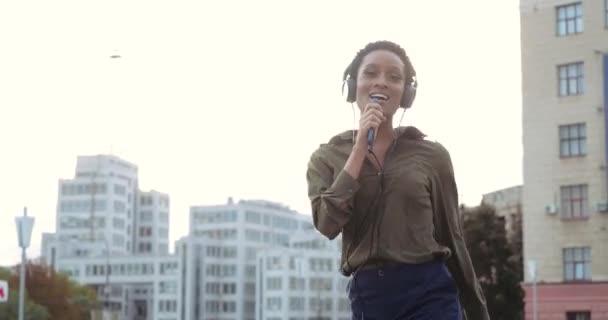 Νέοι μοντέρνα Αφρικής φοιτητής ξοδεύει ελεύθερο χρόνο με τα πόδια γύρω από την πόλη με ακουστικά, τραγουδά τραγούδια σε έξυπνο τηλέφωνο, όπως το μικρόφωνο, απολαμβάνει τον ήχο, χορεύει ανέμελα, απολαμβάνει το Σαββατοκύριακο, μπροστά γυρίσματα — Αρχείο Βίντεο