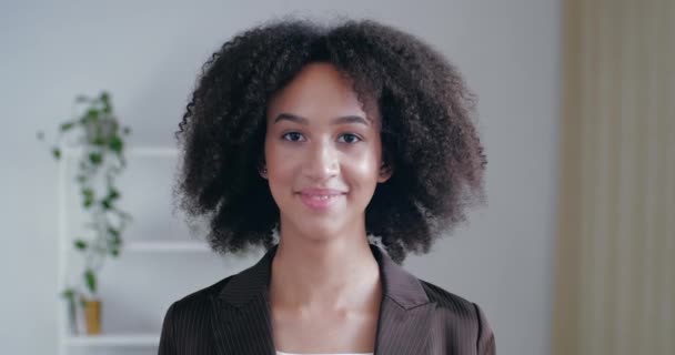 Jovem americana de cabelos encaracolados está no escritório em casa no quarto na frente da câmera, sorri amigavelmente amplamente com dentes brancos, olha alegremente afetuosamente, conceito de atratividade, coração aberto — Vídeo de Stock