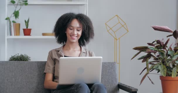 Kobieta rasy mieszanej dorosłych, używająca laptopa do wideokonferencji w pracy, odpowiadająca na pytania przez telefon internetowy. African lady vlogger macha ręką, komunikując się z przyjaciółmi przez kamerę internetową, siedzi w domu na kanapie — Wideo stockowe