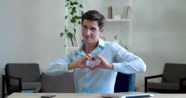Genç çekici iş adamı ofisinde oturuyor, elleriyle parmaklarını kalp şeklinde kıvırıyor, sevgi göstergesi, romantik bir sempati sembolü çiziyor, öpücükler gönderiyor.