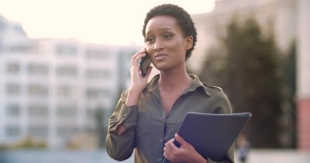 Successful American business woman stoi na zewnątrz na parkingu, posiada folder z dokumentami umowy, omawia negocjacje przez telefon, dialog z klientem, używa smartfona do komunikacji podczas przerwy — Wideo stockowe