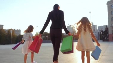 Genç anne esmer kadın şık bir iş elbisesi içinde yürüyor, iki kız arkadaşının elini tutuyor, mağazadan alınmış beyaz bebek elbiseleri, parlak alışveriş çantaları, aile alışverişçilerinin dikiz görüntüsü.