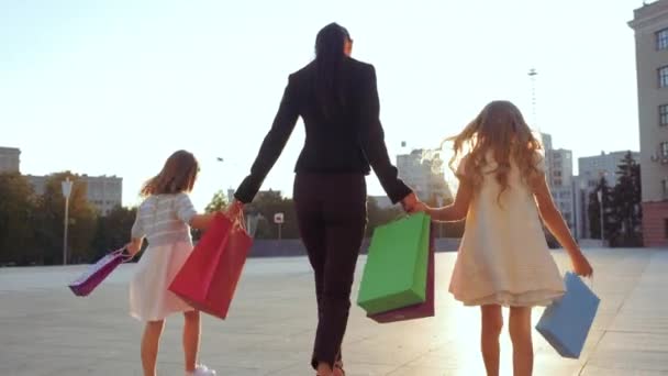 Joven mamá morena esposa camina en elegante traje negro de negocios, toma de la mano de dos hijas novia en nuevos vestidos de bebé blancos comprados en la tienda, lleva bolsas de compras brillantes, familia compradores vista trasera — Vídeos de Stock