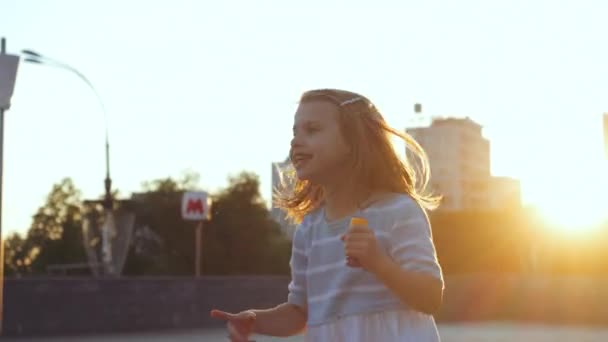 Una niña preescolar activa camina pasa tiempo libre en la calle, salta con alegría, mira hacia otro lado, muestra dirección a lado con el dedo, sostiene burbujas de jabón soplando mirando a la cámara, concepto de la infancia — Vídeo de stock