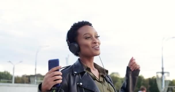 Aktywny model afrykański nosi słuchawki skórzana kurtka tańczy idąc ulicą do muzyki, śpiewa piosenki, przenosi rękę do melodii, trzyma nowoczesne urządzenie smartfona, lubi słuchać ze słuchawkami — Wideo stockowe