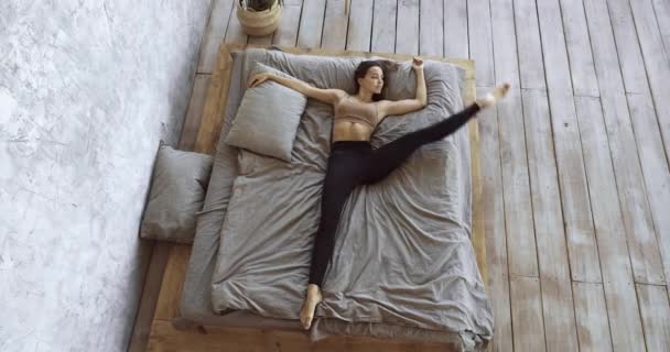 Joven gimnasta femenina se despierta por la mañana en casa haciendo ejercicios de cintura, entrenar acostada en la cama en una habitación acogedora, estirando los músculos de la espalda y las piernas, preparando el cuerpo para un día largo, yoga en posición horizontal — Vídeos de Stock