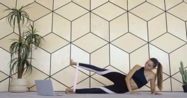 Karantina zamanında spor giyinen genç bir kadın, ev içinde, siyah spor giyim egzersizleri yapıyor. Bayanın bacağını elastik bantla kaldırmasına konsantre olun. Envanter ve spor konseptiyle gergin kaslar.