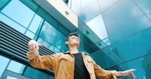 Gros plan élégant attrayant mec adolescent homme porte une veste à la mode et des lunettes de soleil, danse sur la rue mouvements modernes, tourne à la main, se déplace souple avec souplesse, s'amuse en plein air près du bâtiment urbain en verre — Video