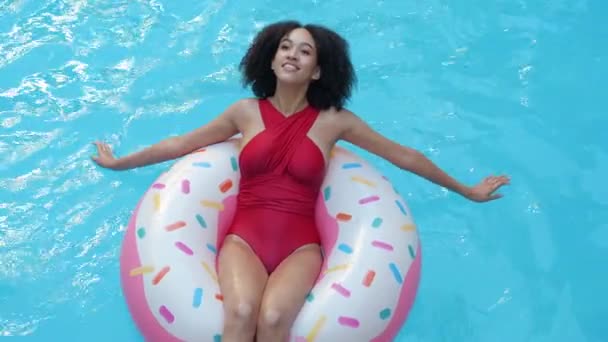 Африканская модель американка сидит в надувном пончике, гребля режет голубую воду в бассейне руками, загорает, улыбается, смотрит в камеру, закрывает глаза, расслабляется, смотрит сверху — стоковое видео