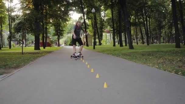 Freestyle Rolschaatsen bij zonsondergang. Jonge langharige man met baard rolschaatser danst tussen kegels in een leuke avond in een stadspark met duim omhoog. — Stockvideo