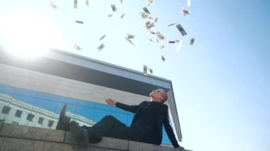 Başarılı genç iş adamı, kazanan açık havada mavi cam binanın yanında duruyor, şık bir siyah takım elbise giyiyor, havaya para atıyor, ikramiye kazanıyor, dolar dağıtıyor, başarı kavramı, alttaki görüş.