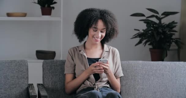 Улыбающаяся афро-американская женщина тысячелетия держит смартфон в руке, наблюдая за видео из социальных сетей, сидящим на диване в гостиной. Счастливая дама смешанной расы, пользующаяся мобильными приложениями, смеясь смотрит в камеру — стоковое видео