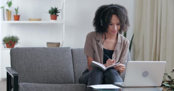 Afrykański student uczy się w domu zdalnie w kwarantannie siedząc na kanapie z laptopem, rozmawiając na konferencji online z nauczycielem, słuchając kurs webinarny, robiąc notatki, słuchawki mikrofon — Wideo stockowe