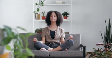 Sakin, rahat bir Afrikalı kadın evinde, gözleri kapalı, bacak bacak bacak üstüne atmış, meditasyon yapıyor, yoga yapıyor. Karışık ırk kızı evde bitkilerle derin derin nefes alma alıştırması yapıyor.