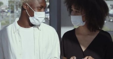 Afro-Amerikalı genç kız ve melez etnik erkek yan yana duruyor, koruyucu tıbbi maskeler takıyor, virüs enfeksiyonunu önlüyor, internette yazı yazıyor, diyalog kuruyor.