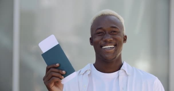 Молодий афроамериканець в білому одязі посміхається, радісно відкриває губи з щастям, показує квитки на літак і паспорт до камери, святкує свята. — стокове відео