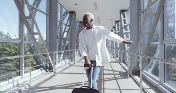 Aktiver afroamerikanischer Typ ausländischer Student trägt weißes Hemd, tanzt am Flughafen mit Koffer, bewegt aktiv seinen männlichen Körper fröhlich, genießt den Urlaub, wartet am Flughafenterminal auf das Flugzeug — Stockvideo