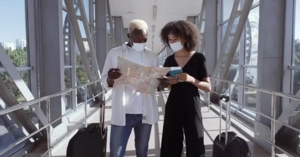 Een paar jonge Afrikanen, vriend met vrouw op het vliegveld, staande met bagage, medische maskers op het gezicht, op zoek naar een landkaart, op zoek naar een uitvalsbasis in nieuw land, huwelijksreis concept — Stockvideo