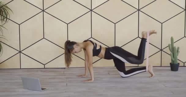 Jonge sportieve vrouw die thuis hippe oefeningen doet, een been schudt met elastische band voor fitness, traint in online trainingslessen op netwerklaptop, vorm behoudt in quarantaine — Stockvideo