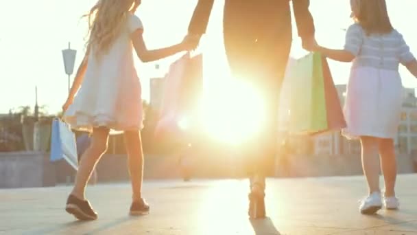 Gros plan des jambes humaines, deux petites filles marchant dans la rue dans des chaussures élégantes, tenant la main des mamans. Mère en talons de luxe à la mode va avec les filles du magasin de shopping, porte en paquets cadeaux — Video