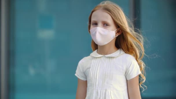 Poważna młoda dziewczyna z długimi złotymi blond lokami nosi białą sukienkę i różową maskę ochronną, stoi na zewnątrz przed kamerą, wyglądając na zdenerwowaną oburzeniem, pandemicznym wirusem epidemii — Wideo stockowe
