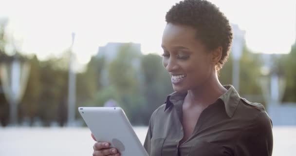 Молодая этническая женщина носит рубашку, стоит на улице на фоне заката, использует компьютерный планшет для общения, говорит онлайн-звонок на конференцию веб-камеры, активно жестикулирует руками эмоционально — стоковое видео