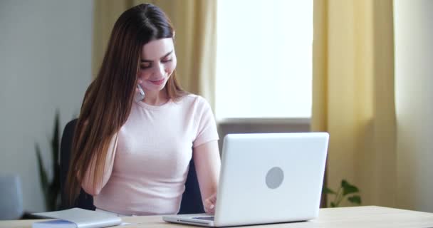 Retrato de mulher jovem com cabelos longos senta-se em casa no escritório na mesa de trabalho, fala por telefone para dispositivo on-line, responde a perguntas, tipos no laptop, faz compras, conversa com amigos na rede de smartphones — Vídeo de Stock