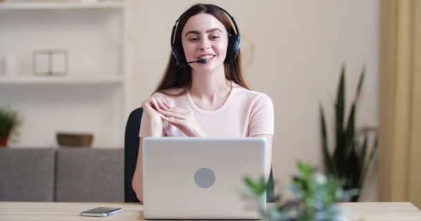 Retrato de jovem bonito atraente mulher chamada gerente consultor sentado à mesa em casa no escritório, conversando com clientes on-line em microondas, usando fones de ouvido, comunicando ativamente gesticule — Vídeo de Stock