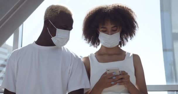 Пара афроамериканцев, двое молодых подростков, носят защитные медицинские маски на лицах, стоят вместе, разговаривают, смотрят на смартфон, общаются через интернет в сети по телефону, концепция карантина — стоковое видео
