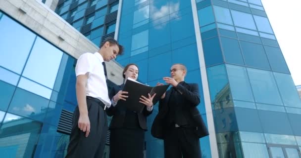 Grupo de jóvenes empresarios juntos en la calle contra el fondo de la construcción de rascacielos se comunica, discute caso, hombre tiene carpeta con documentos en sus manos y muestra colegas de acuerdo — Vídeos de Stock