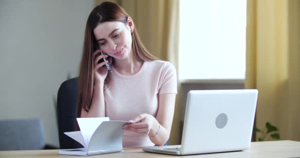 Freelancer mulher senta-se à mesa no escritório fala no telefone, folheando páginas de caderno, conversando no smartphone durante a pausa no trabalho. Menina se comunica na conexão celular em casa — Vídeo de Stock