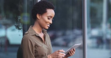 Afro-Amerikalı bir kadın cam binanın yanında oturuyor, elinde bilgisayar tableti tutuyor, internetten bilgi topluyor, parmaklarını iyi gösteren kameraya bakıyor, rıza sembolü, tamam mı?
