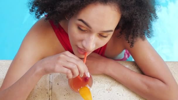 Ripresa ravvicinata di sofisticata donna afroamericana soddisfatta seduta in acqua blu vicino a bordo piscina, bere cocktail analcolico attraverso il bar esterno di paglia nel complesso turistico dell'hotel, vista dall'alto — Video Stock