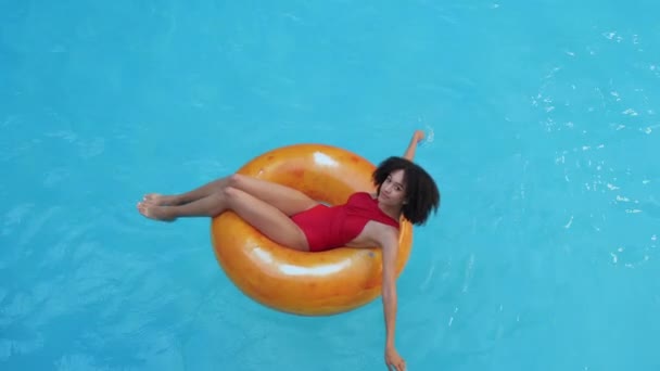 Afro american curly-haired model woman resting, lies with closed eyes in orange inflatable swimming lifebuoy, floats on water in pool, sunbathes thin beautiful body under summer sunlight, top view — Stock Video