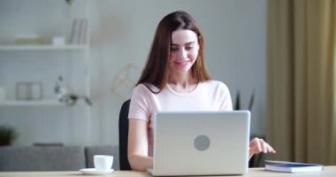 Genç, gülümseyen bir kadın masada oturuyor, bilgisayar ekranına bakıyor, öğretmenle video görüşmesi yapıyor, internetten e-öğrenme üzerine çalışıyor, not defterine bakıyor, notlar okuyor, kurantina...
